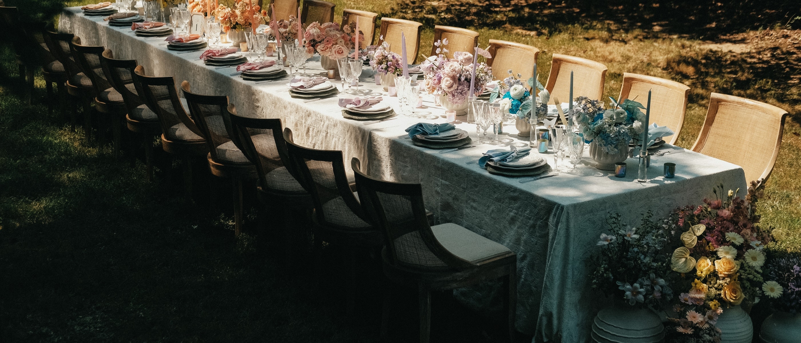 Long garden dinner table dressed in linen, glassware, and pastel florals under dappled light
