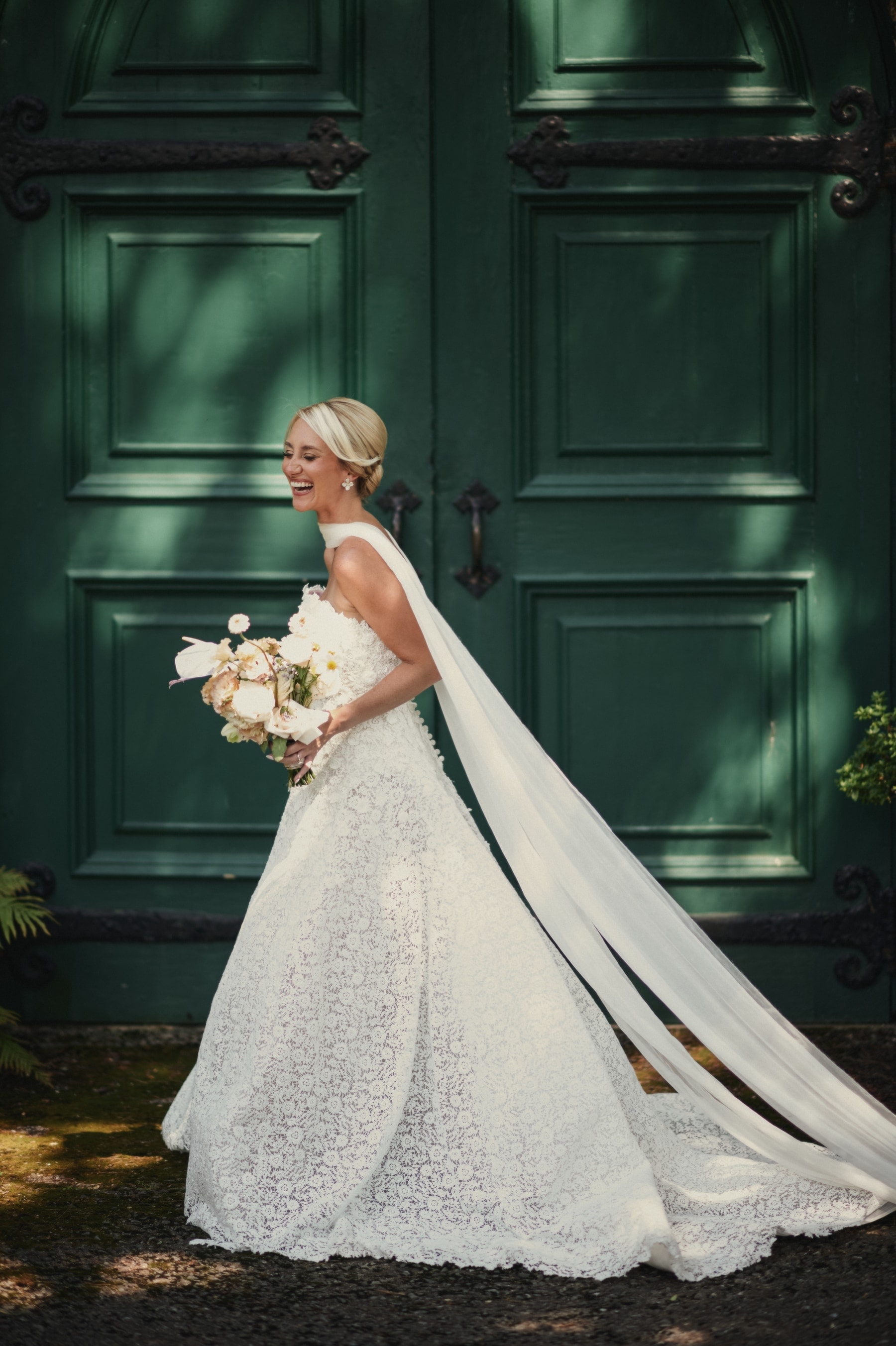 Bride laughing with her bouquet before deep-green chapel doors, veil falling to the ground