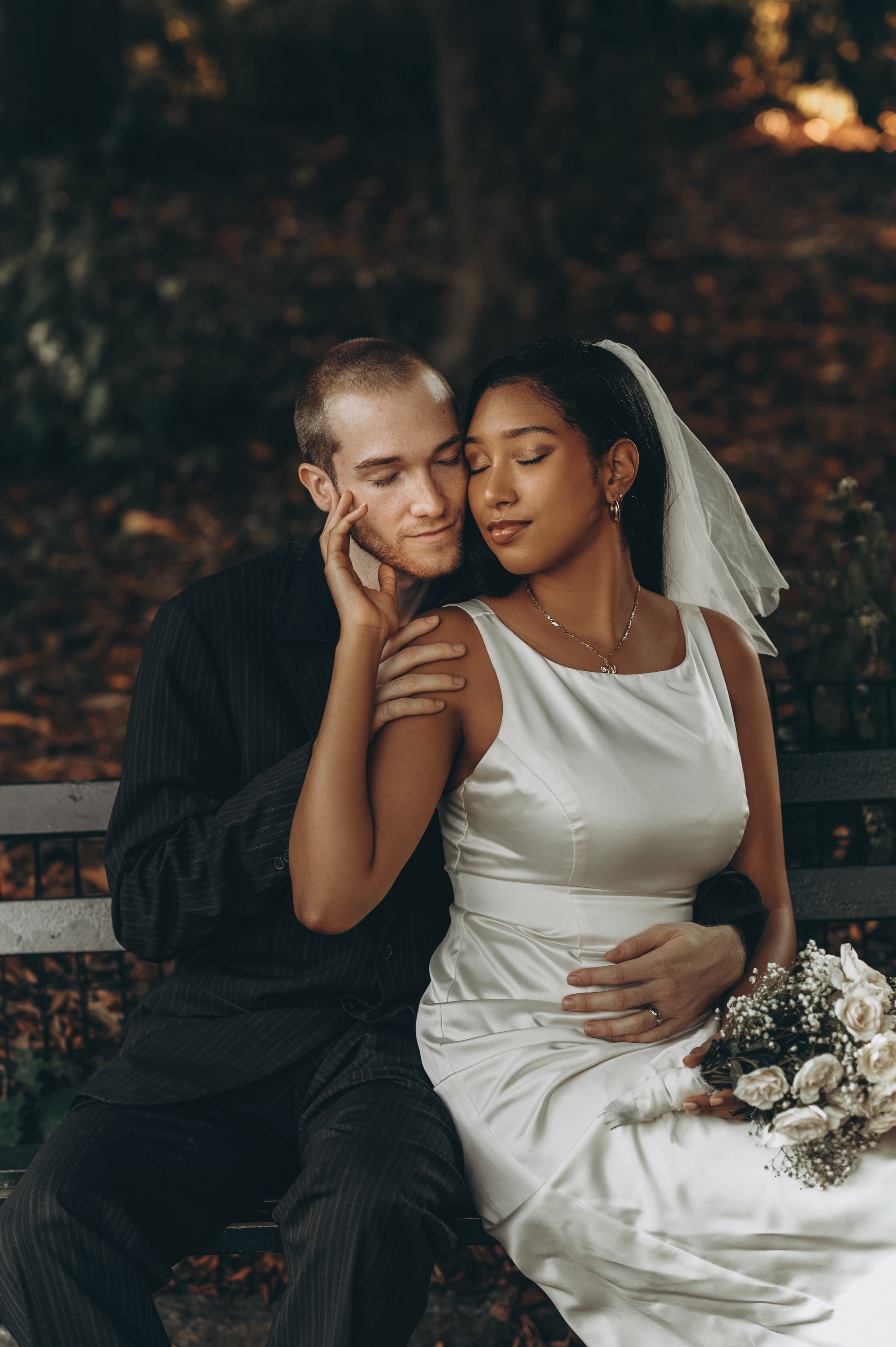 Bride and groom resting cheek to cheek on a park bench in autumn light, eyes closed
