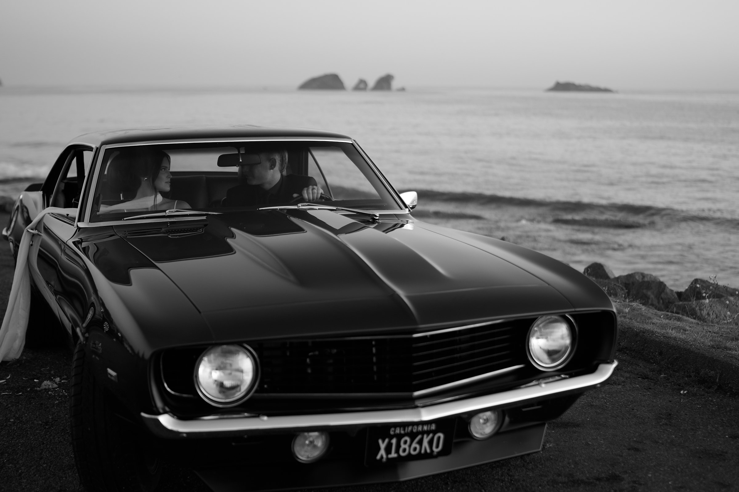 Bride and groom in a vintage Camaro parked by the sea, black and white