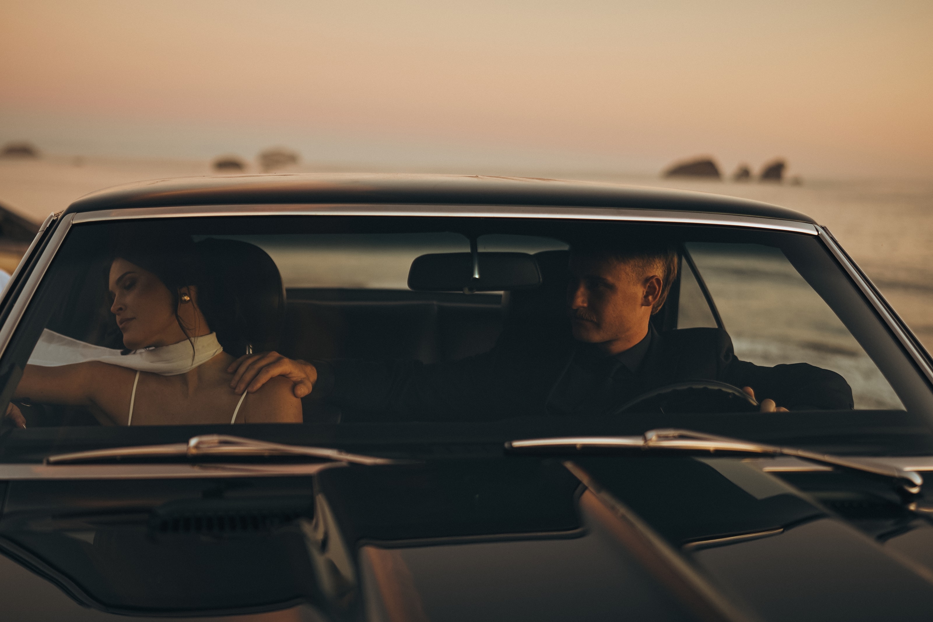Bride and groom in a vintage car at dusk on the coast, her veil trailing from the window — fine art film photograph