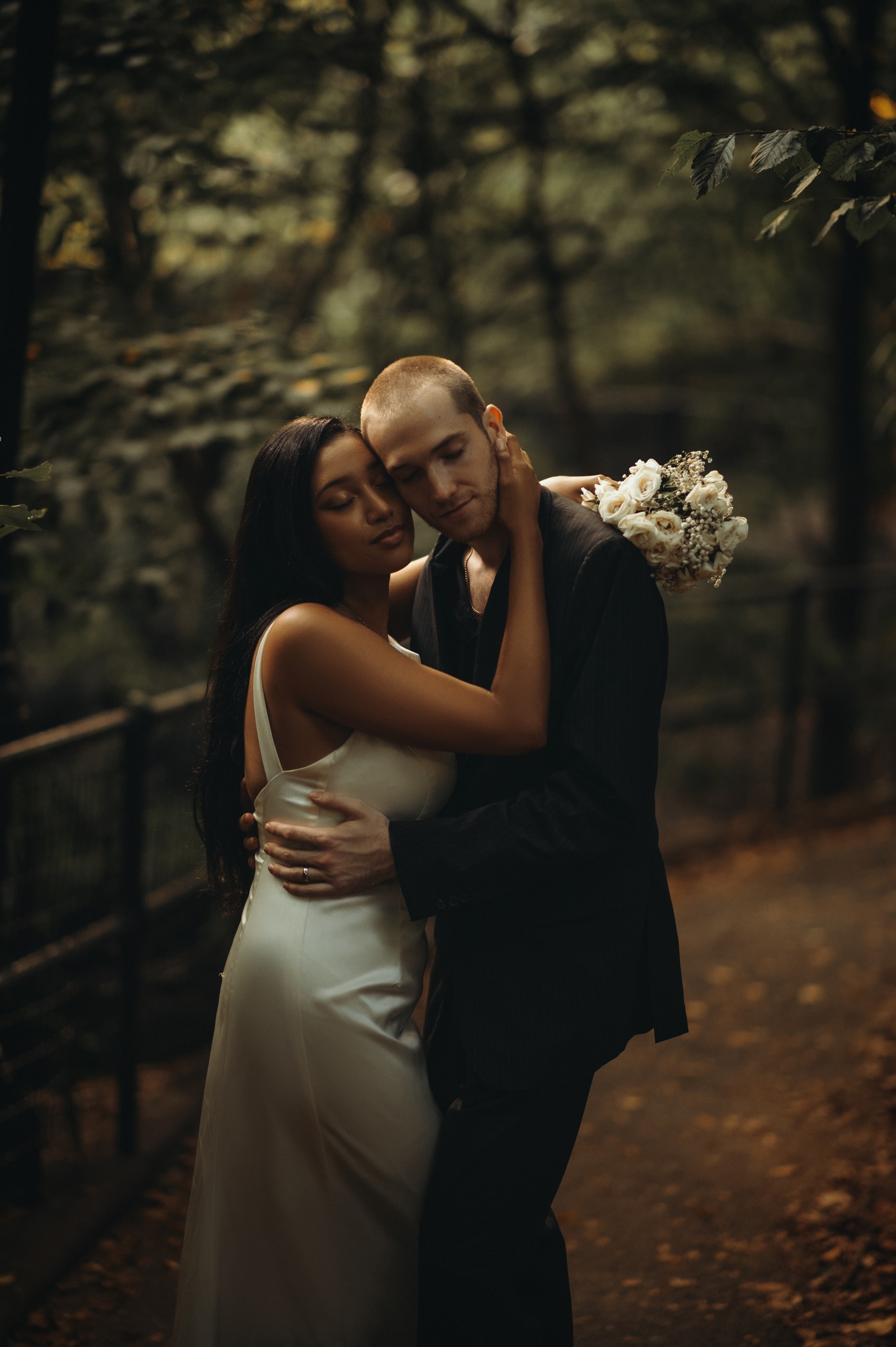 June holding Elias on a wooded park path, bouquet of white flowers over his shoulder