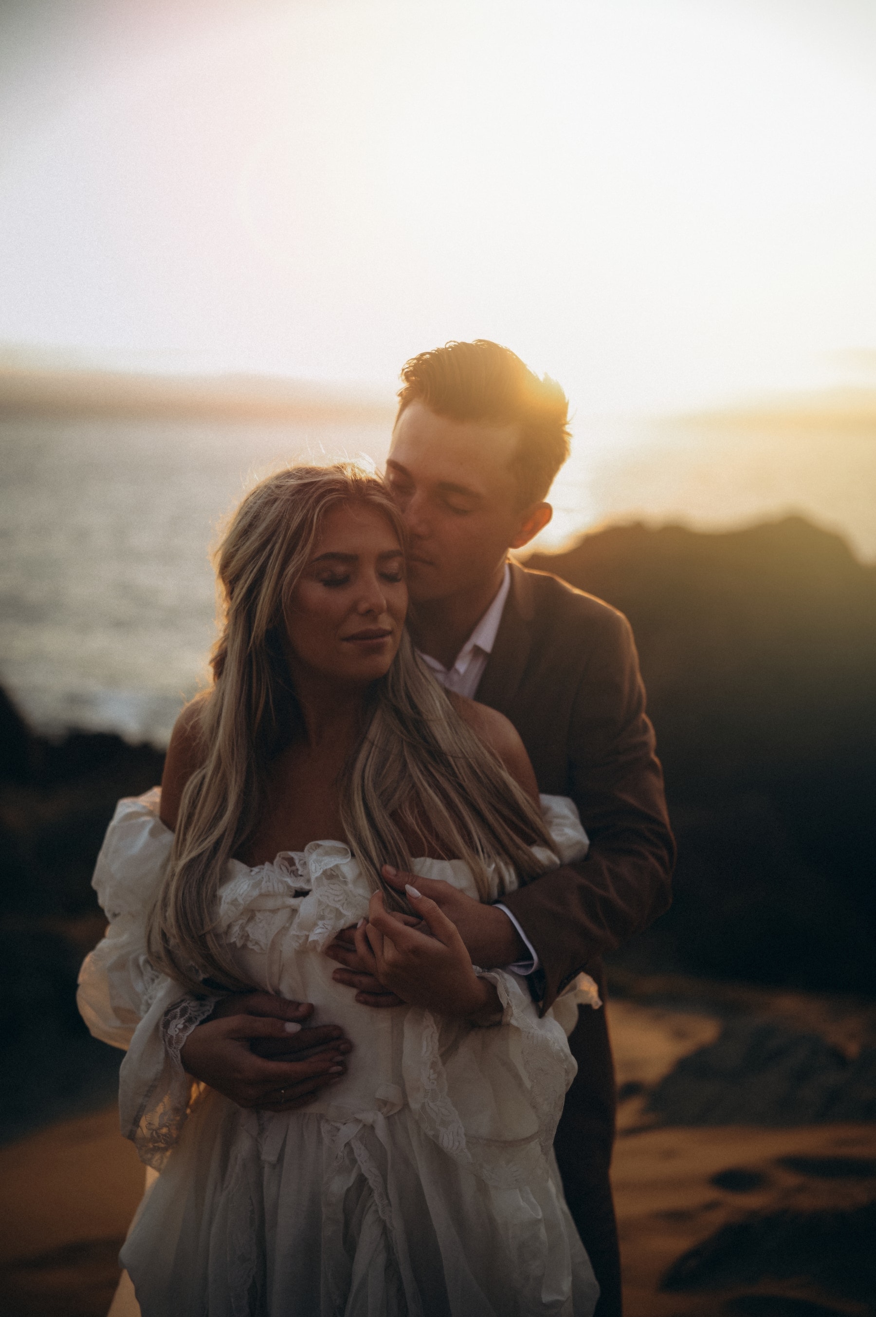 Groom holding the bride on a coastal bluff at sunset, gown in the wind