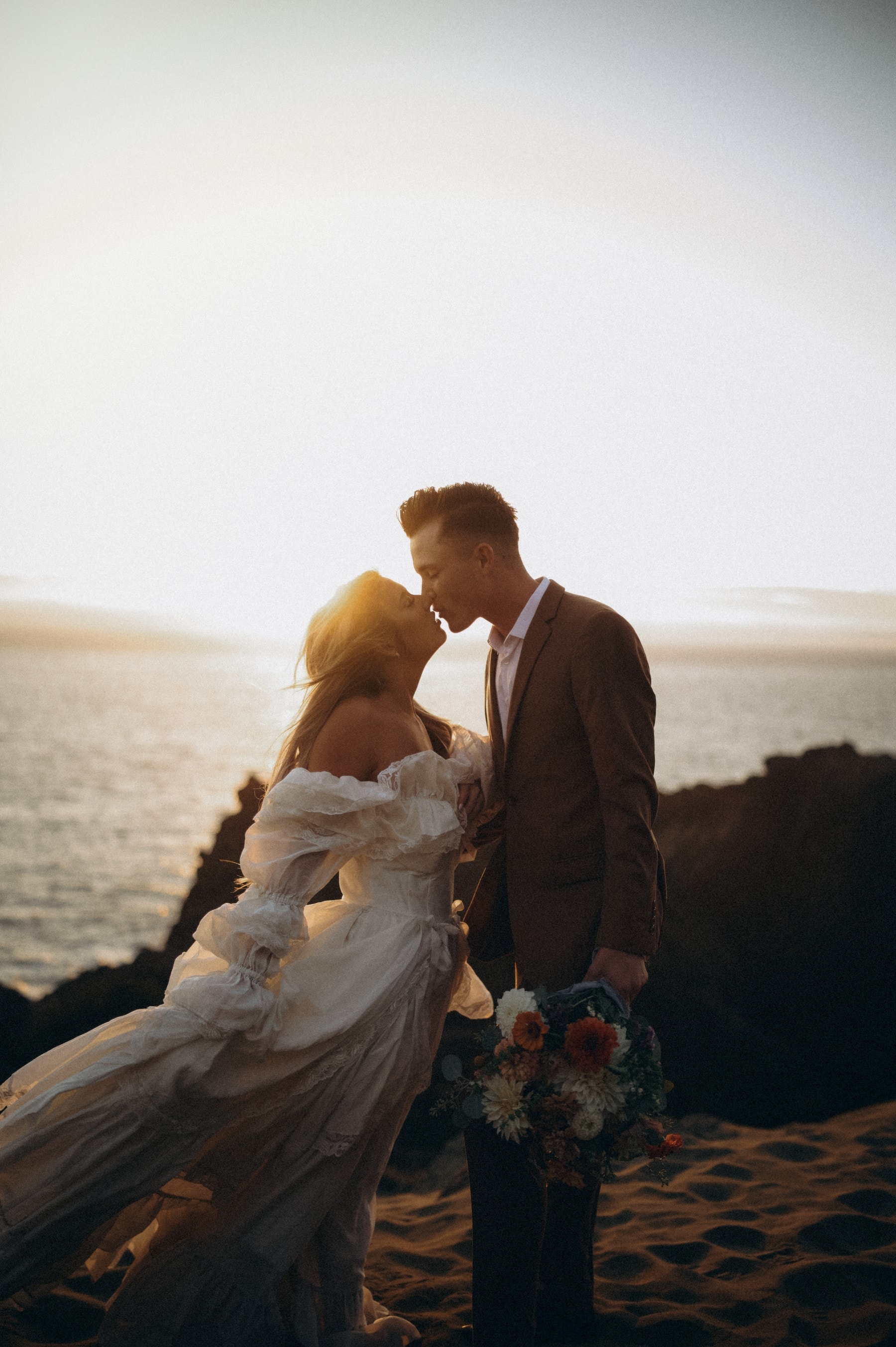 Blake and Maria kissing at sunset on a coastal cliff, backlit by golden light