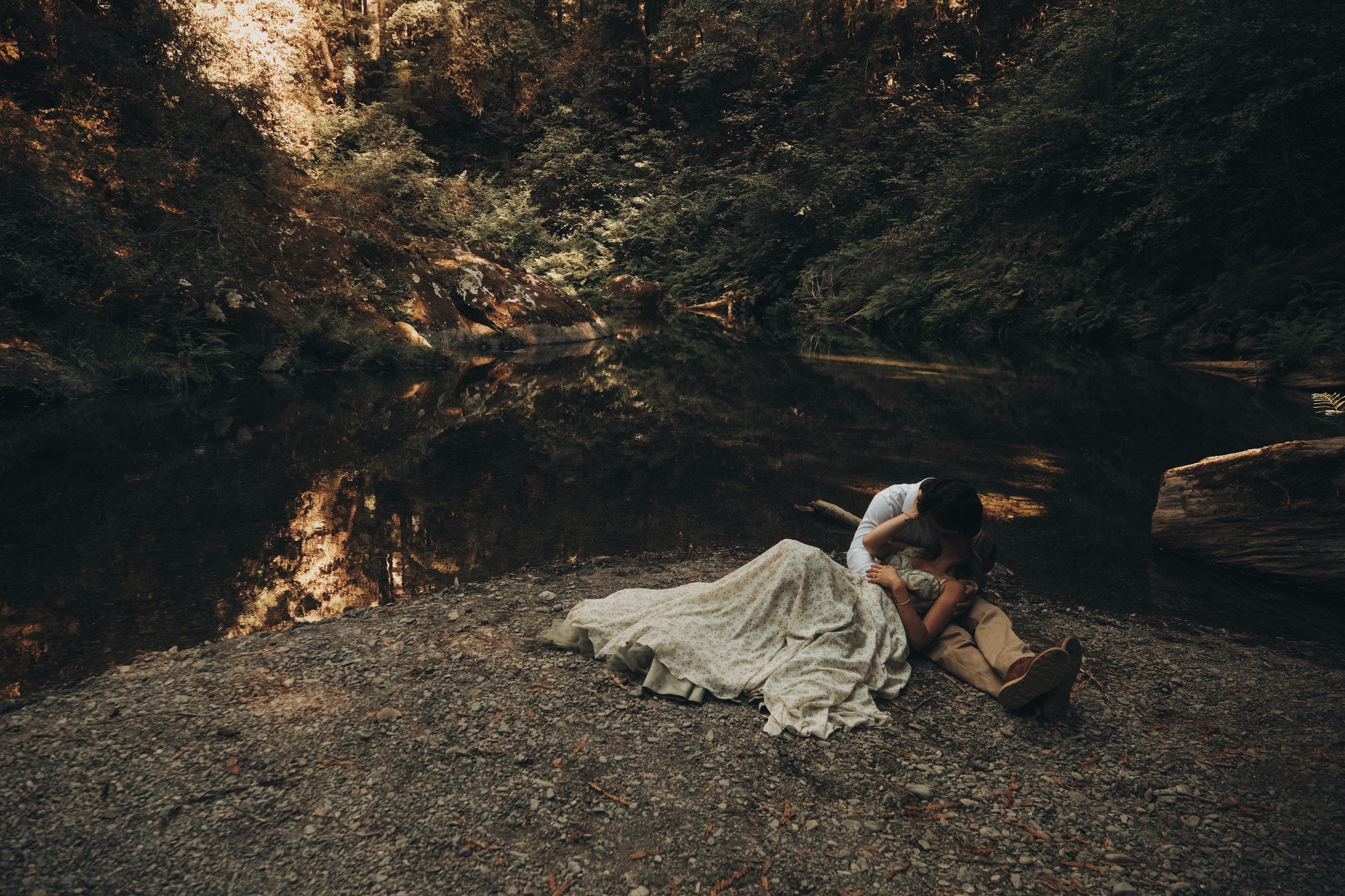 Blake and Maria lying beside a dark forest river beneath the redwoods, her gown across the stones